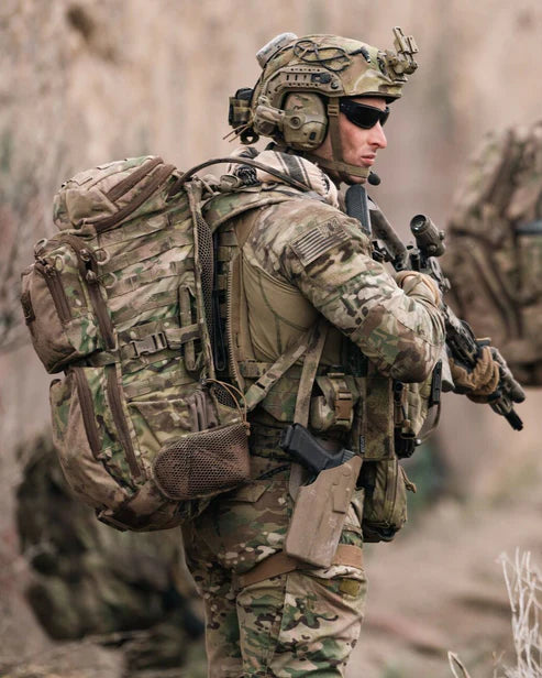 Ein Soldat in voller Tarnkleidung, Helm und Sonnenbrille steht mit taktischer Ausrüstung und einem Gewehr im Freien und trägt den EBERLESTOCK FAC Track - Coyote Brown Rucksack mit MOLLE-System vor einem unscharfen, erdigen Hintergrund.