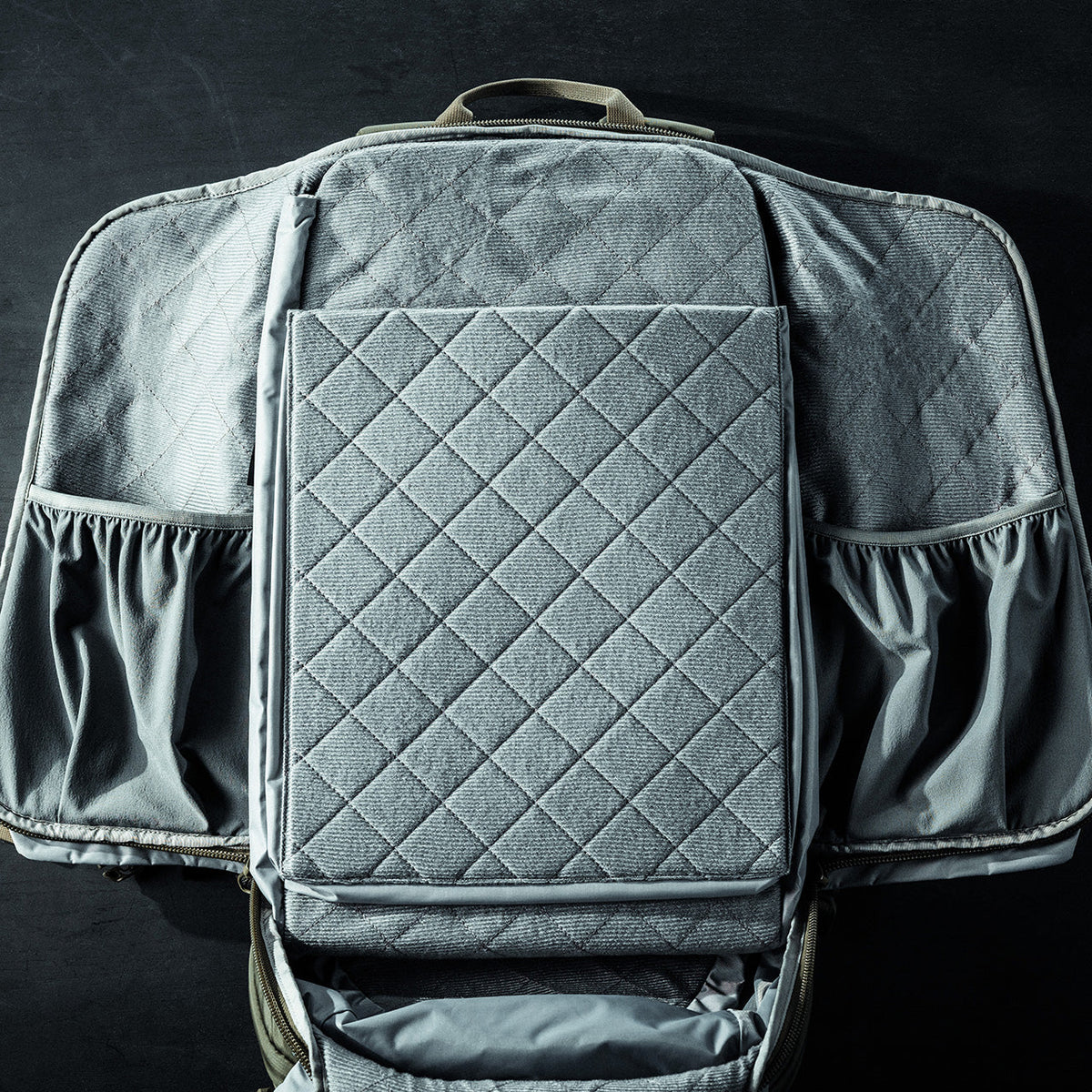 Eine graue, gesteppte, rechteckige Wickelunterlage befindet sich in einem offenen EBERLESTOCK Eberlestock Fade Adapt Rucksack mit weichen, elastischen Seitentaschen, die alle auf einer dunklen Oberfläche ausgestellt sind.