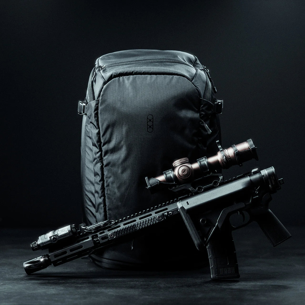 Ein schwarzer EBERLESTOCK Fade Adapt Rucksack steht aufrecht auf einer dunklen Fläche, an die ein modernes schwarzes Gewehr mit Zielfernrohr und Taschenlampe gelehnt ist. Die Szene hat einen minimalistischen, dunklen Hintergrund.