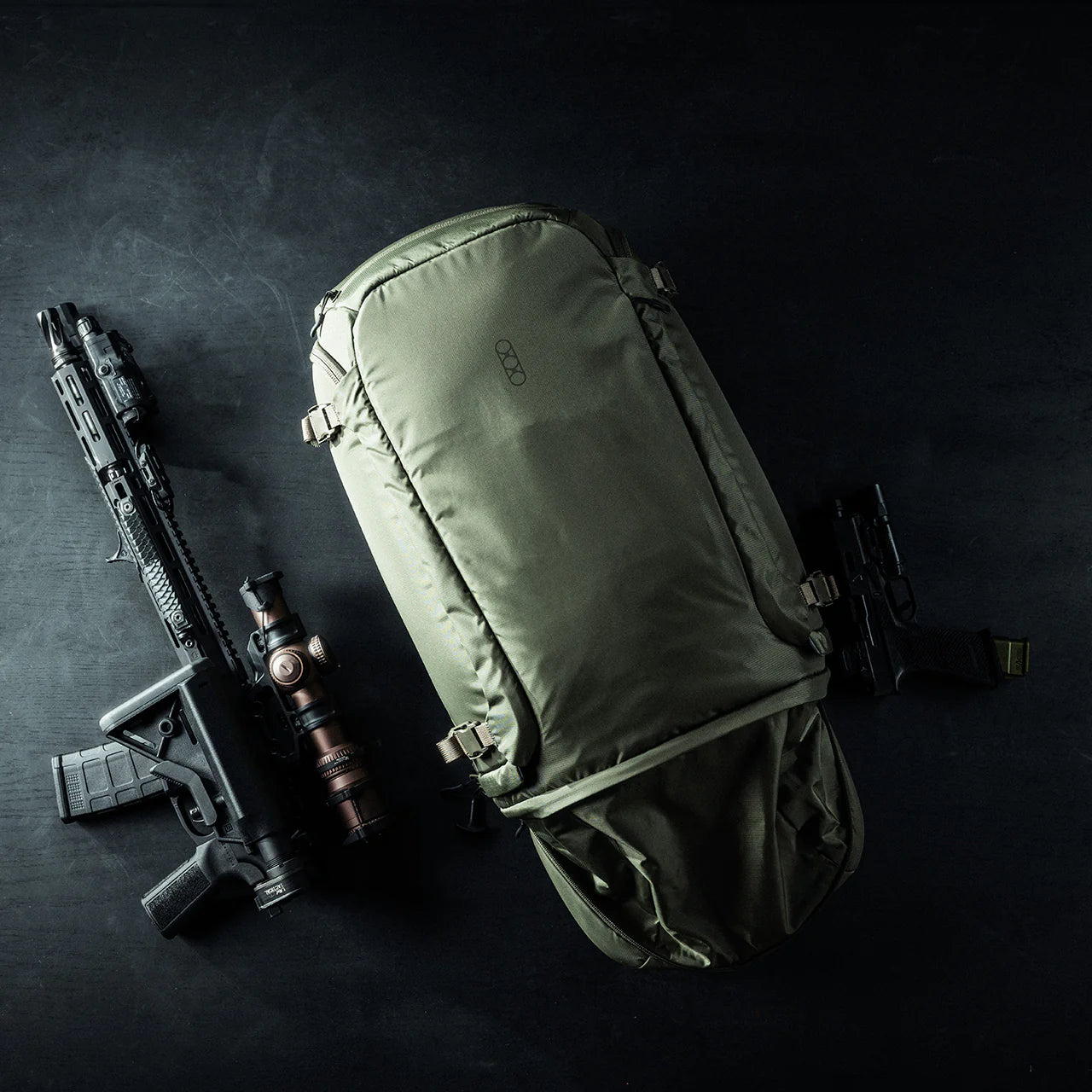 Ein olivgrüner EBERLESTOCK Fade Adapt taktischer Rucksack mit ausklappbarer Scheide ruht auf einer dunklen Fläche, flankiert von einem schwarzen Gewehr mit Zielfernrohr auf der linken und einer schwarzen Handfeuerwaffe auf der rechten Seite, die seine vielseitige Funktionalität demonstrieren.