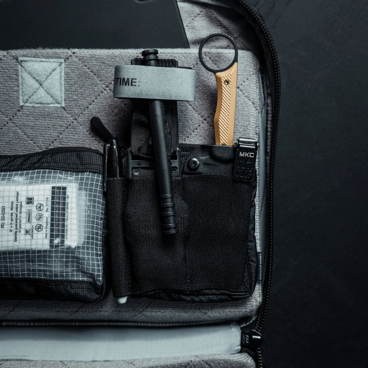Eine Nahaufnahme der EBERLESTOCK Fade Admin Pouch Small aus Ripstop-Nylon, in der Tourniquet, Kugelschreiber, Taschenlampe, Multitool und medizinisches Zubehör in organisierten MOLLE-kompatiblen Fächern untergebracht sind.