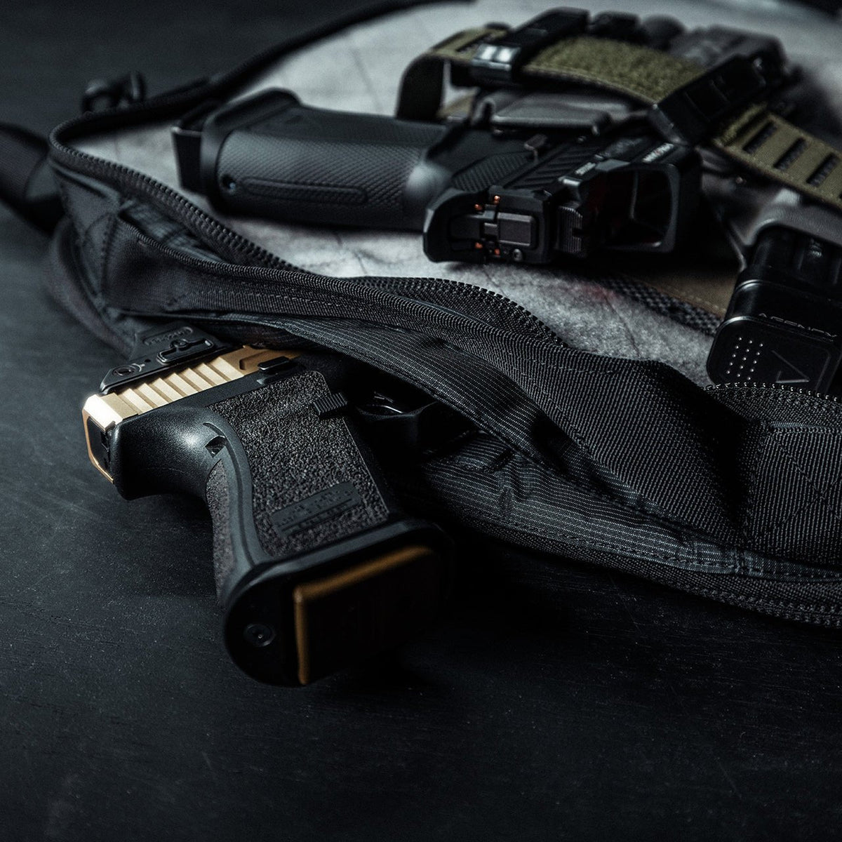 Eine goldfarbene Handfeuerwaffe, ein Magazin und eine taktische Ausrüstung kommen teilweise aus einer geöffneten EBERLESTOCK Eberlestock Fade Brief auf einer dunklen Oberfläche zum Vorschein - Accessoires, die üblicherweise mit der zivilen Polizeiarbeit verbunden sind.