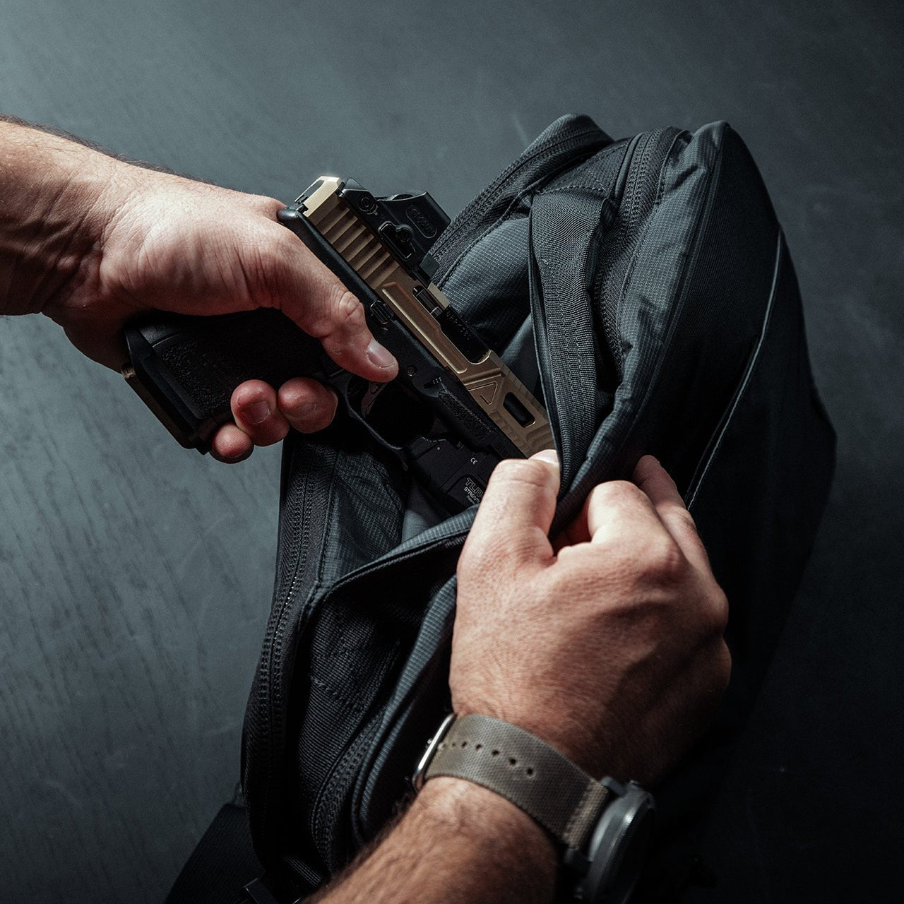 Man sieht Hände, die eine halbautomatische Handfeuerwaffe aus der Vordertasche einer EBERLESTOCK Eberlestock Fade Brief nehmen, vor einem dunklen Hintergrund, der an zivile Polizeiarbeit denken lässt.