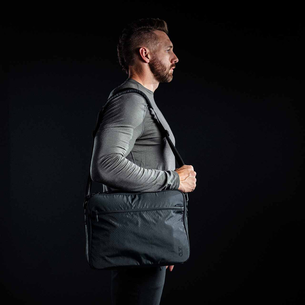 Ein Mann in einem grauen langärmeligen Hemd steht im Profil vor einem schwarzen Hintergrund und trägt die EBERLESTOCK Eberlestock Fade Brief Crossbody.