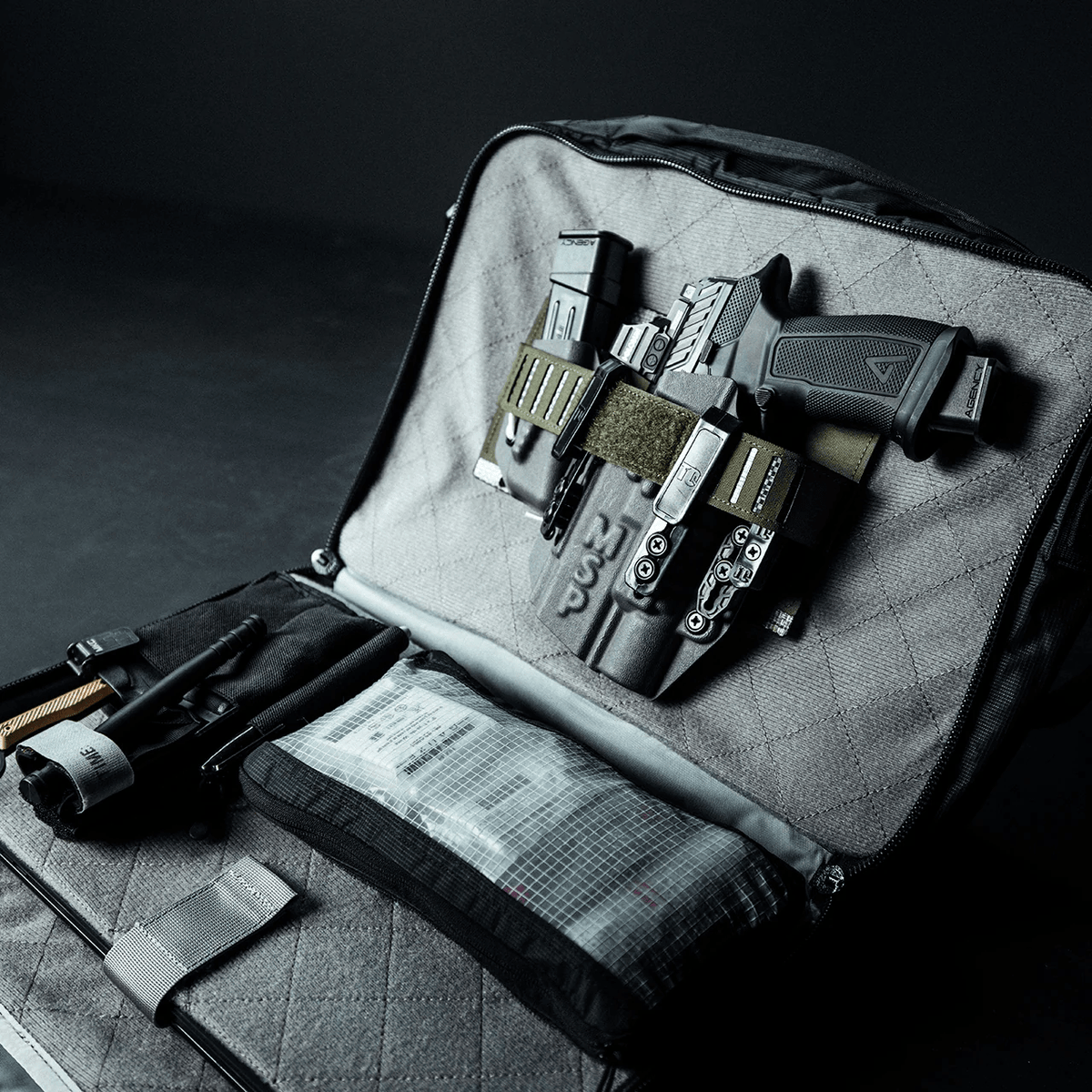 Ein geöffneter EBERLESTOCK Eberlestock Fade Brief zeigt eine Handfeuerwaffe mit Holster, zusätzliche Magazine, eine Taschenlampe und ein Erste-Hilfe-Set - ideal für die zivile Polizeiarbeit vor einem dunklen Hintergrund.