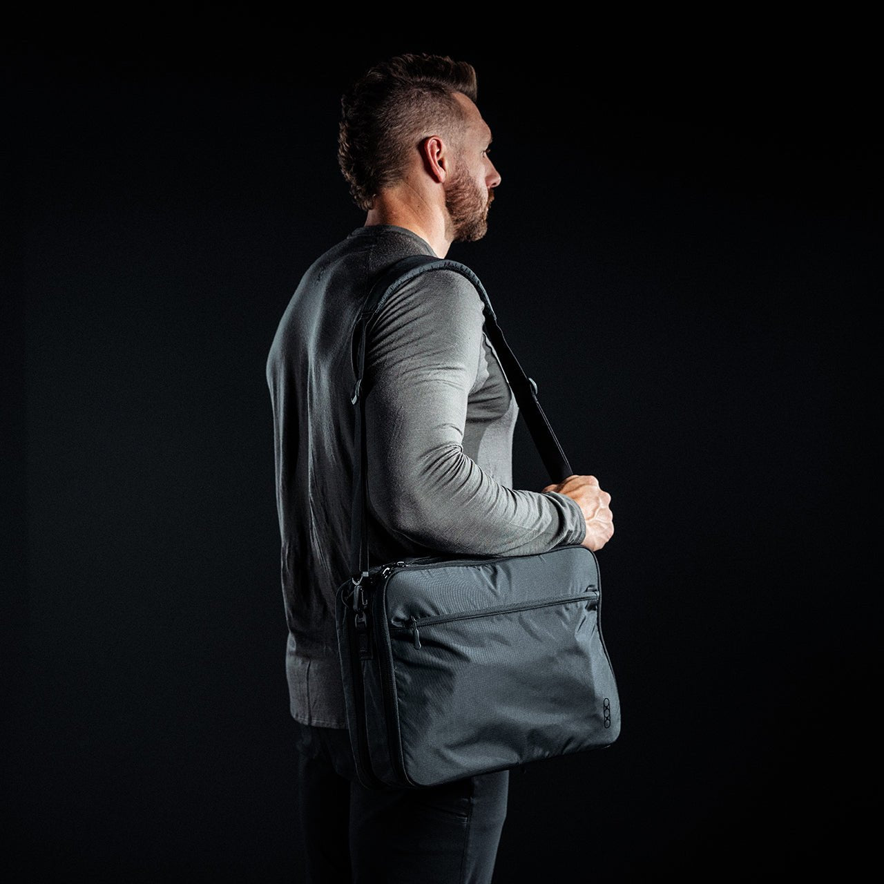 Ein Mann mit kurzen Haaren und Bart, der ein graues, langärmeliges Hemd trägt, steht vor einem dunklen Hintergrund und hält einen EBERLESTOCK Eberlestock Fade Brief über seine Schulter.