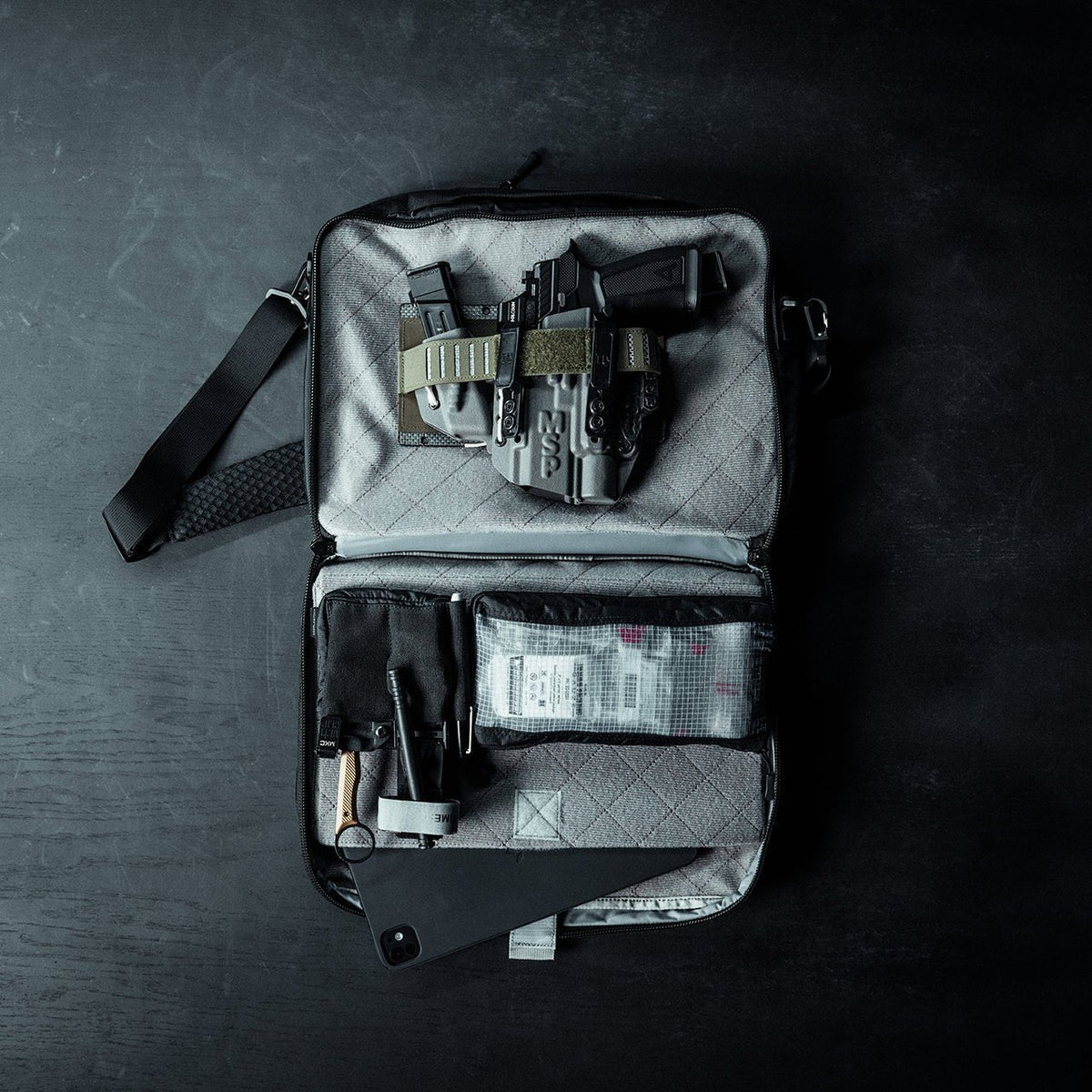 Ein EBERLESTOCK Fade Brief in Schwarz liegt ohne Reißverschluss auf einer dunklen Fläche und zeigt eine Handfeuerwaffe im Holster, Ersatzmagazine und medizinisches Material - kompakte Utensilien für die zivile Polizeiarbeit, alles fein säuberlich in den Fächern angeordnet.