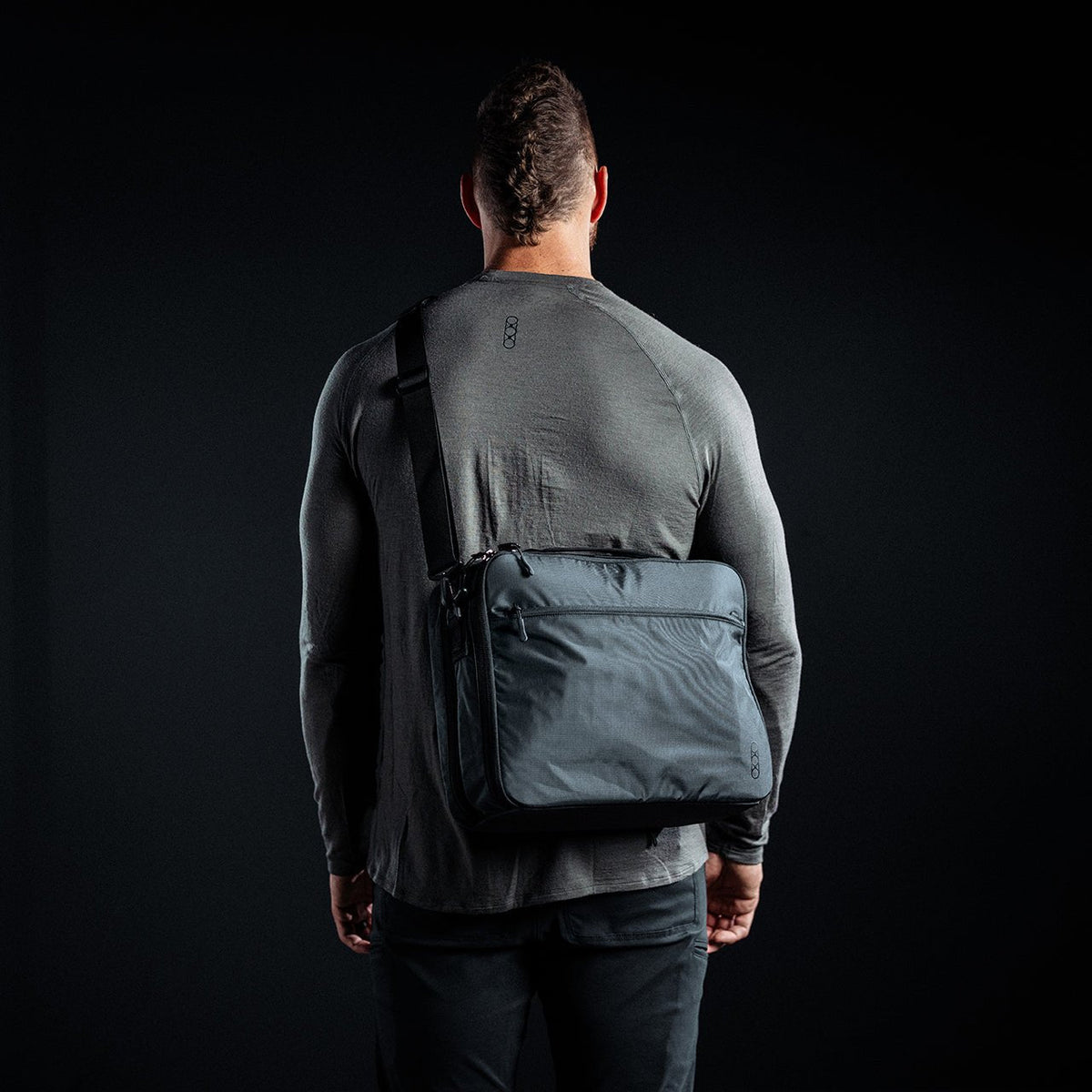 Eine muskulöse Person, bekleidet mit einem taillierten grauen langärmeligen Hemd und einer schwarzen Hose, steht mit dem Rücken zur Kamera vor einem dunklen Hintergrund und trägt die EBERLESTOCK Fade Brief Laptop Aktentasche über einer Schulter.