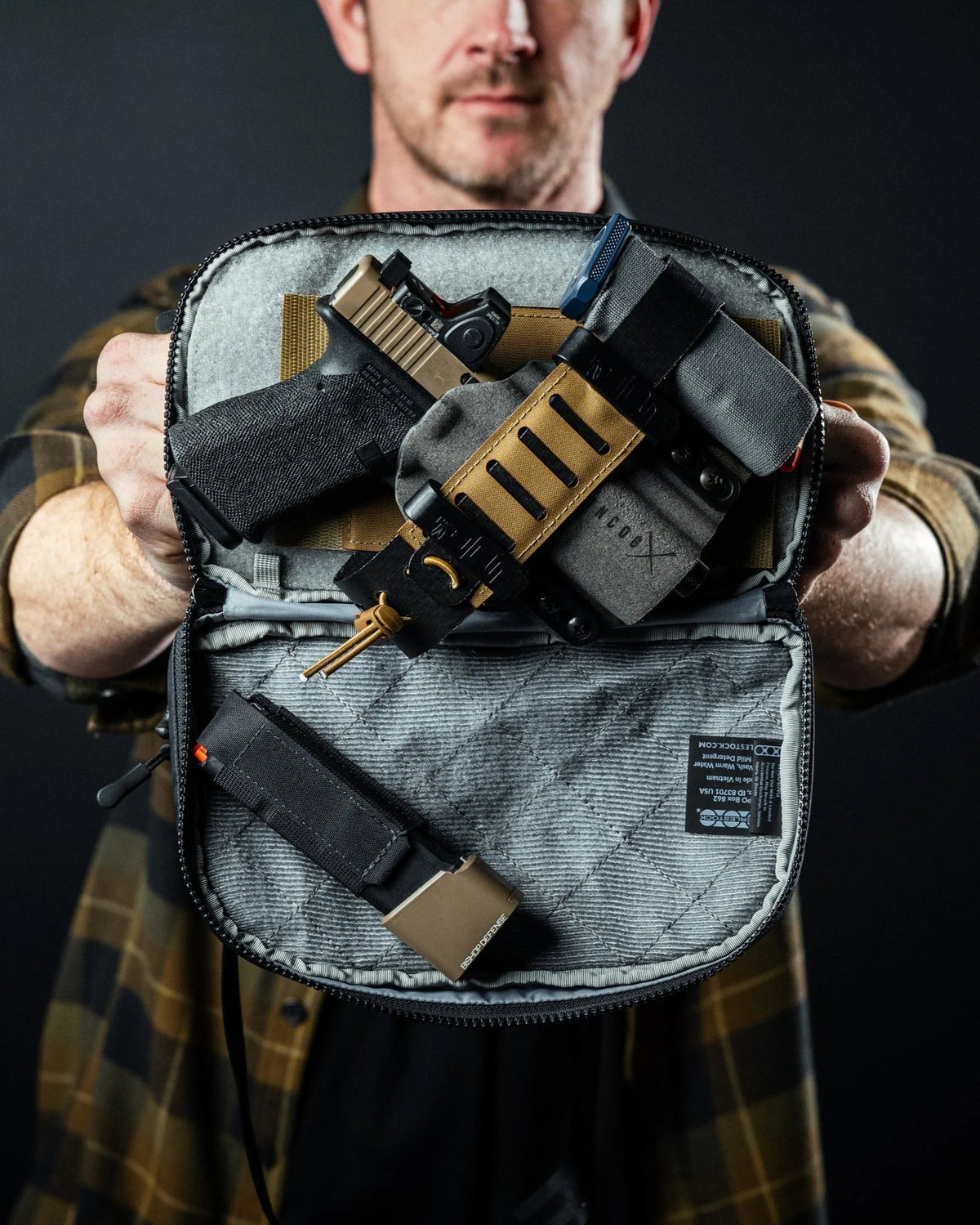 Ein Mann in einem karierten Hemd hält eine geöffnete graue EBERLESTOCK Eberlestock Fade Flex Crossbody-Tasche in die Kamera und zeigt eine Handfeuerwaffe in einem Holster und ein Magazin in der Tasche vor einem dunklen Hintergrund.