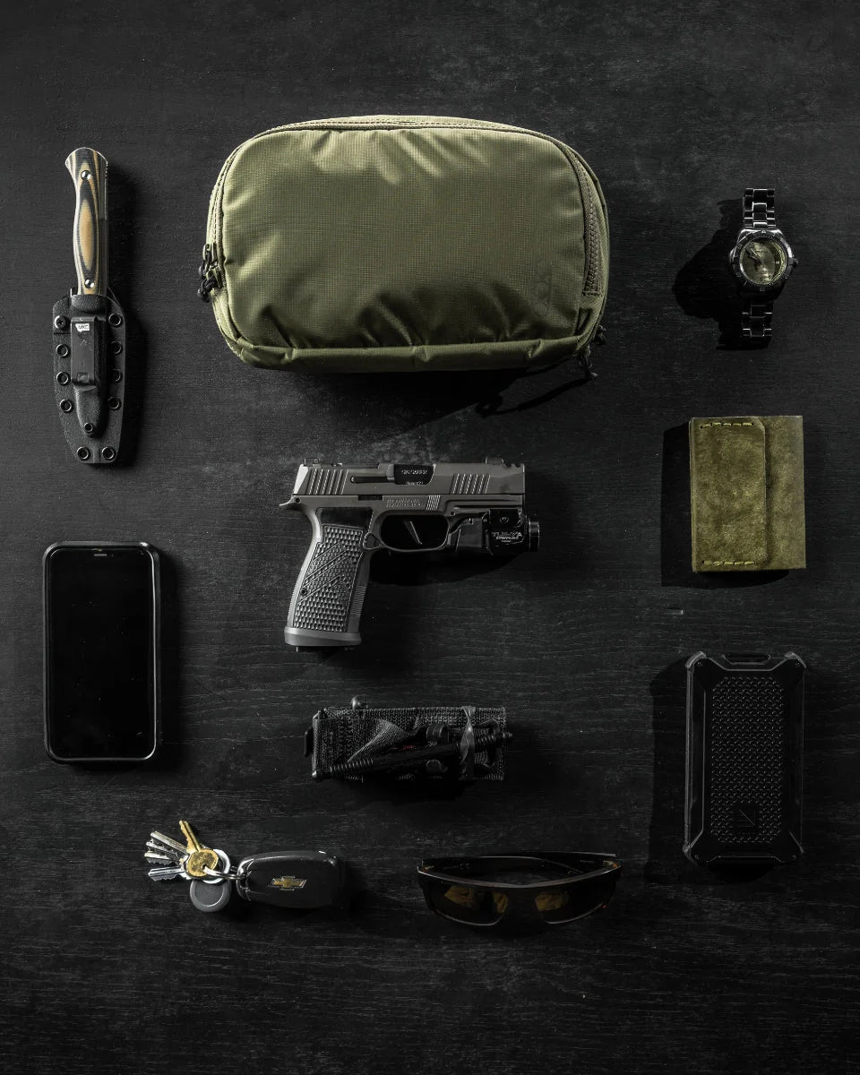 Flaches Foto von Gegenständen des täglichen Bedarfs auf einer dunklen Oberfläche, das den EBERLESTOCK Fade Flex Hüftrucksack in Grün zusammen mit einer Armbanduhr, einem Messer, einer Pistole, einem Smartphone, einer Geldbörse, einem Multitool, einer Sonnenbrille, einem Schlüsselbund mit Schlüsseln und einem kleinen schwarzen Etui zeigt.