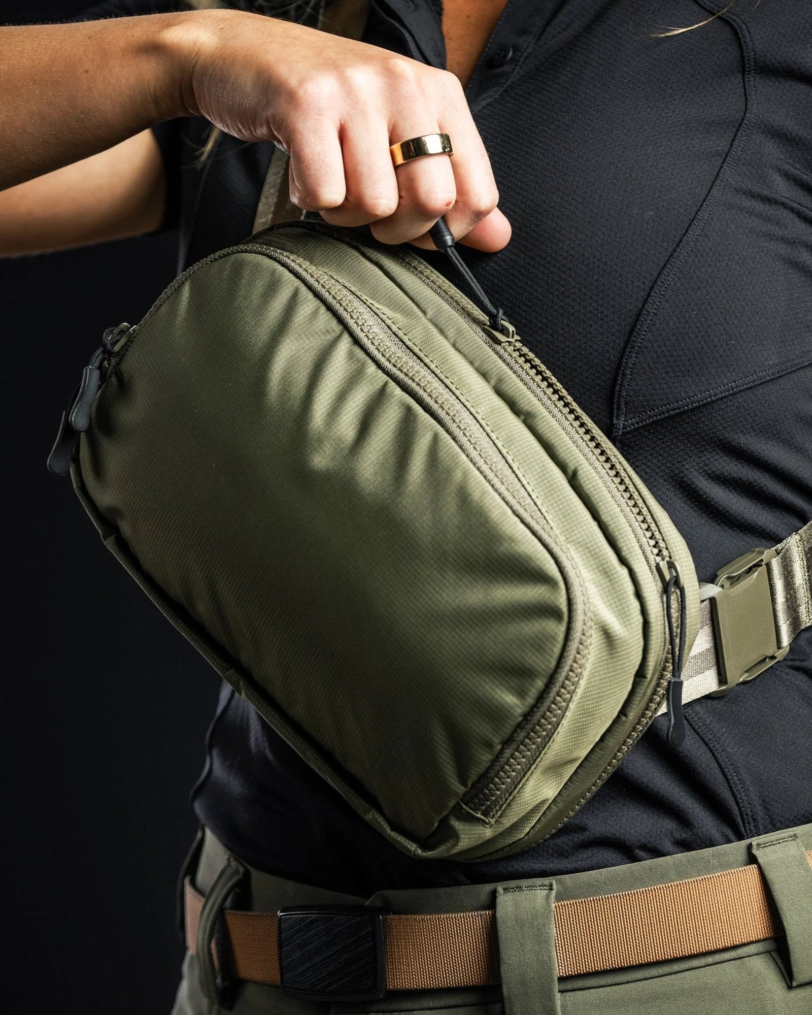 Eine Person mit schwarzem Hemd und hellbraunem Gürtel hält eine olivgrüne EBERLESTOCK Fade Flex Gürteltasche mit Reißverschluss und Riemen.