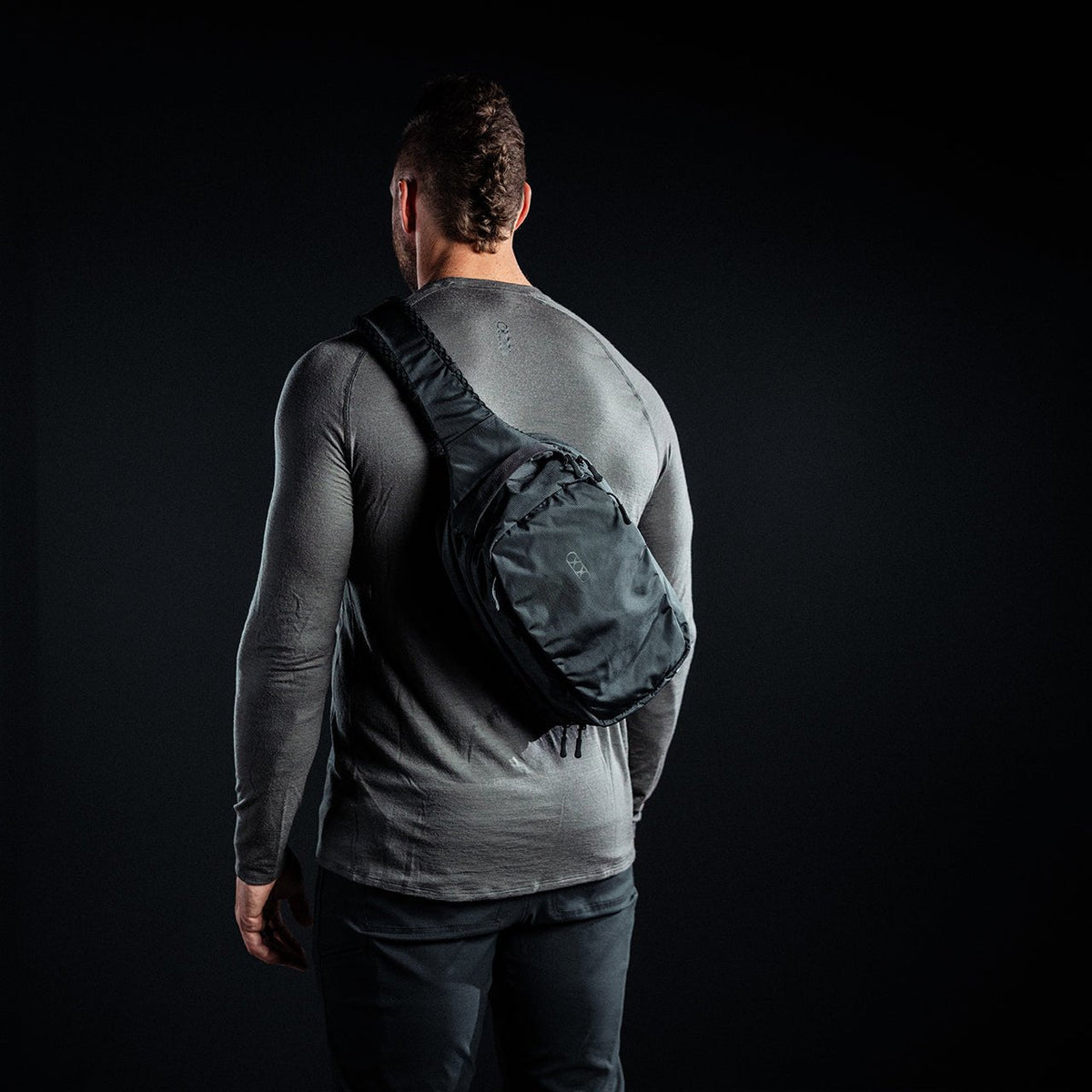 Ein muskulöser Mann, von hinten gesehen, trägt ein graues, langärmeliges Hemd und eine schwarze Hose. Auf seinem Rücken trägt er einen EBERLESTOCK Eberlestock Fade Sling Rucksack, der vor einem dunklen Hintergrund steht.