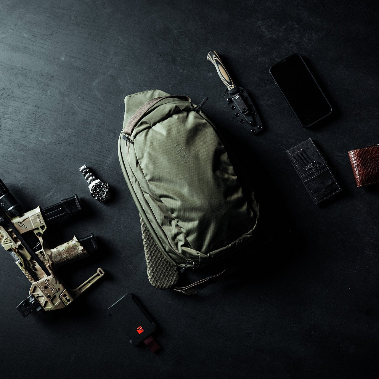 Eine flache Anordnung von Alltagsgegenständen auf einer dunklen Oberfläche, mit dem EBERLESTOCK Fade Sling Rucksack in Olivgrün, einem Messer, einem Smartphone, einer Armbanduhr, einer Brieftasche, einer Taschenlampe, einem Ladegerät und einem Teil eines braunen Gewehrs.
