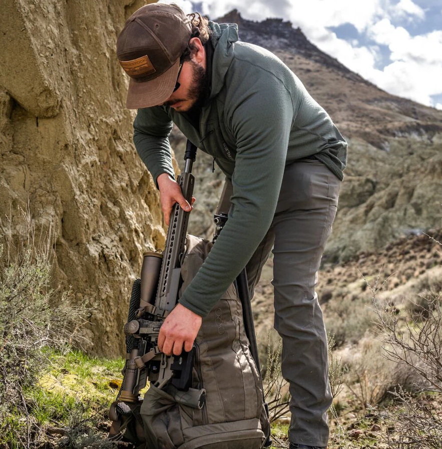Ein Mann in Outdoor-Ausrüstung und mit Mütze packt sein Gewehr mit Zielfernrohr in den EBERLESTOCK FreeFall 2000 Pack. Er steht neben einem felsigen Hügel in den schroffen Bergen und bereitet sich auf sein nächstes Outdoor-Abenteuer vor.