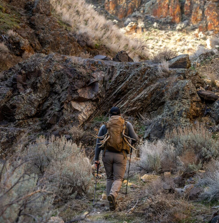 Mit Trekkingstöcken und dem vielseitigen EBERLESTOCK FreeFall 2000 Pack wandert eine Person durch einen felsigen Canyon und trägt auf ihrem Outdoor-Abenteuer Geweihe durch zerklüftetes Gelände und karge Vegetation.