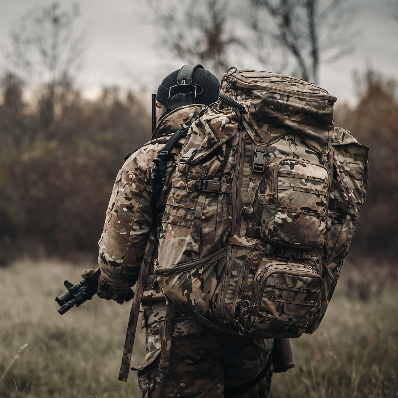 Eine Person in tarnfarbener Militärkleidung mit einem EBERLESTOCK G4 Operator Pack steht im Freien auf einer Wiese, schaut weg und hält eine Schusswaffe. Im Hintergrund sind Bäume und ein bewölkter Himmel zu sehen.
