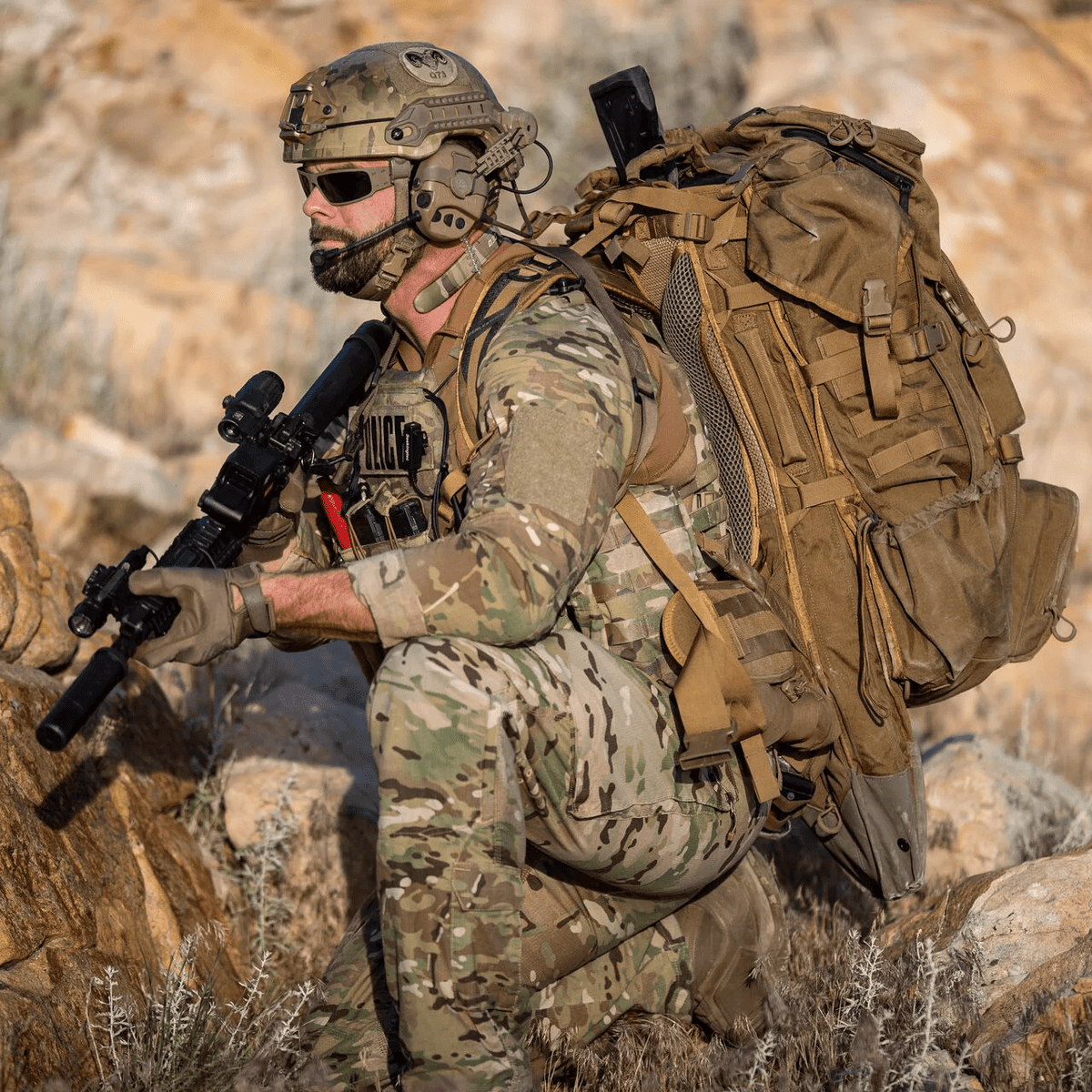 Ein Soldat in Tarnkleidung und mit Helm kniet im Freien mit einem Gewehr, einer Sonnenbrille, einer taktischen POLICE-Weste und einem EBERLESTOCK G4 Operator Pack inmitten von felsigem Gelände und trockenem Gras.