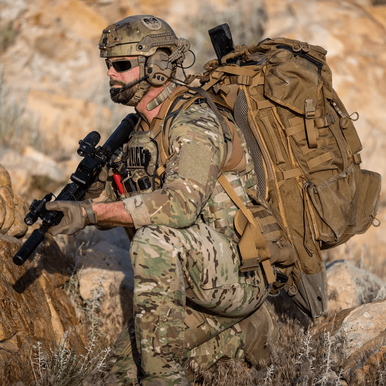 Ein Soldat in Tarnkleidung und mit Helm kniet im Freien mit einem Gewehr, einer Sonnenbrille, einer taktischen POLICE-Weste und einem EBERLESTOCK G4 Operator Pack inmitten von felsigem Gelände und trockenem Gras.