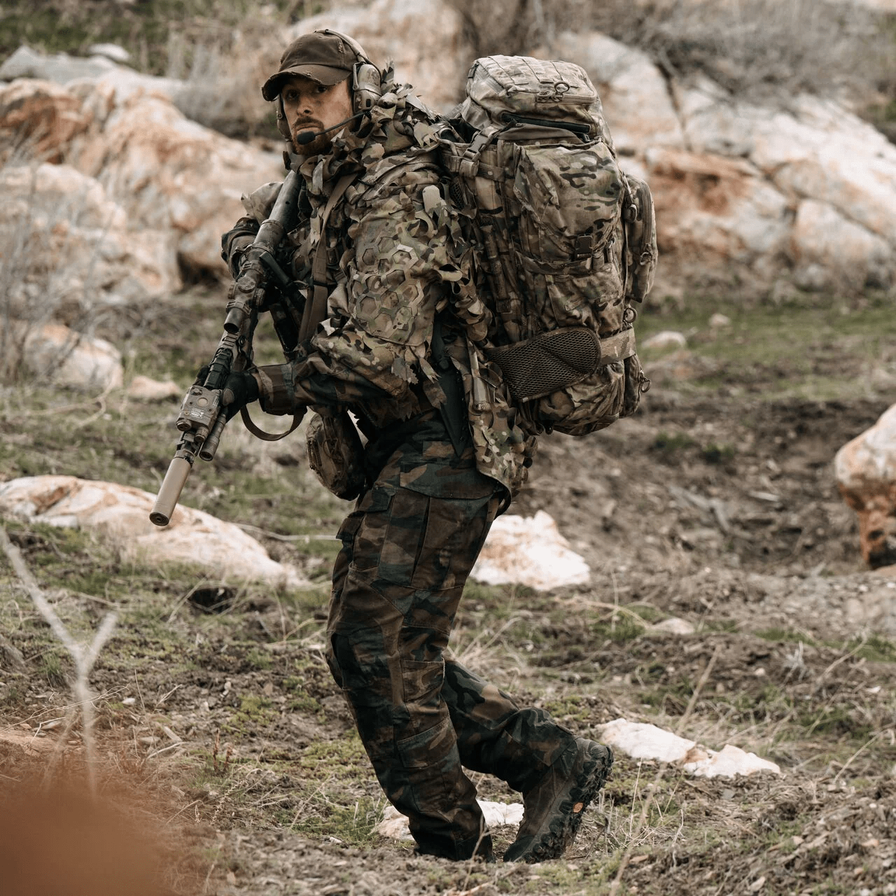 Ein Soldat in Camouflage und Stiefeln durchquert felsiges Gelände und ist mit einem EBERLESTOCK G4 Operator Pack, Mütze, Headset und Gewehr im Anschlag unterwegs.