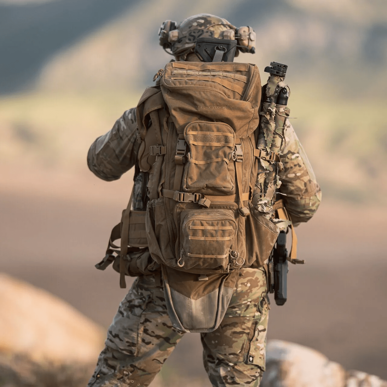 Von hinten gesehen trägt eine Person in voller Tarnkleidung und mit Helm ein EBERLESTOCK G4 Operator Pack und steht im Freien, mit verschwommenen Bergen im Hintergrund.
