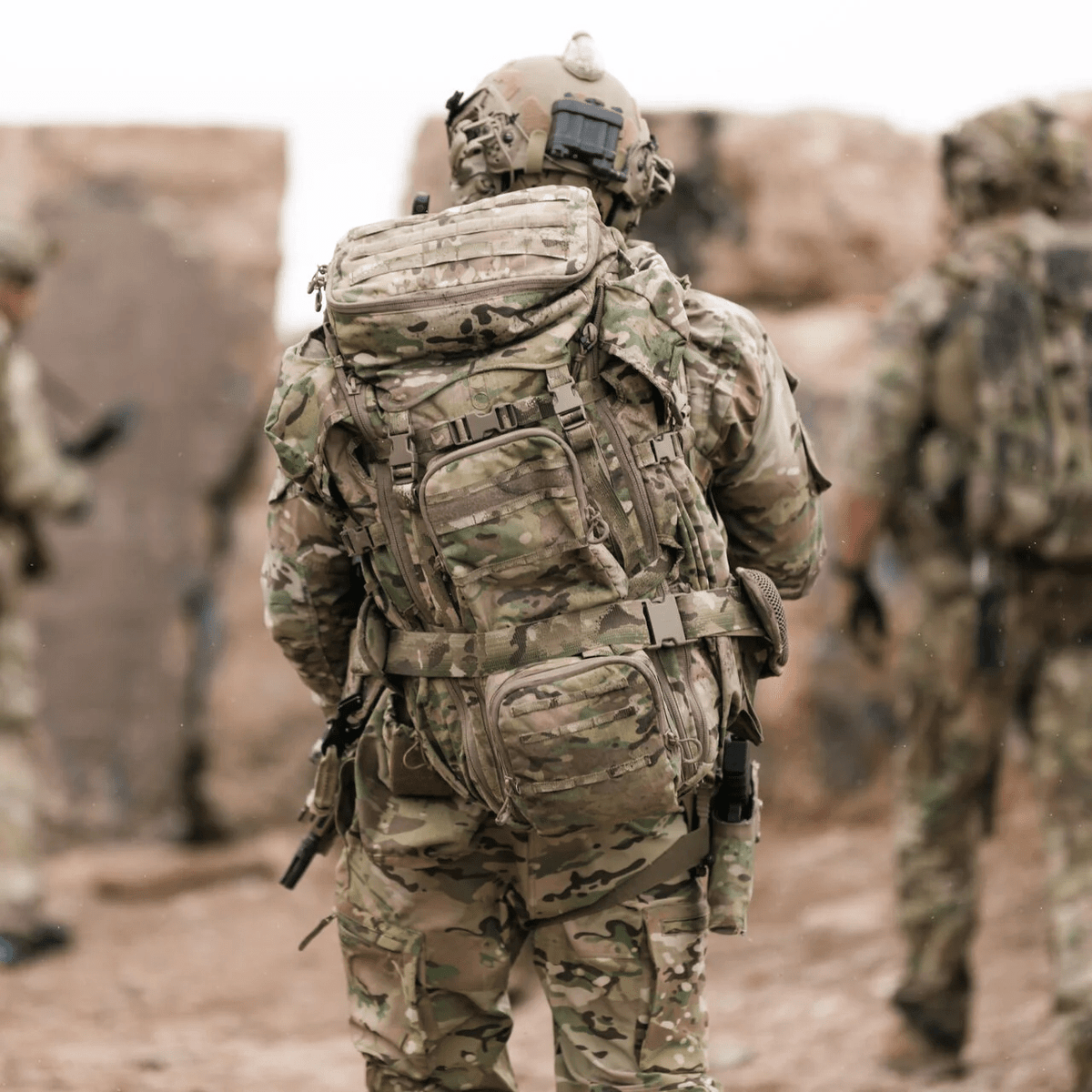 Ein Soldat in voller Tarnkleidung und mit einem EBERLESTOCK G4 Operator Pack steht auf felsigem Untergrund, während zwei weitere Soldaten mit taktischen Rucksäcken im Hintergrund zu sehen sind.