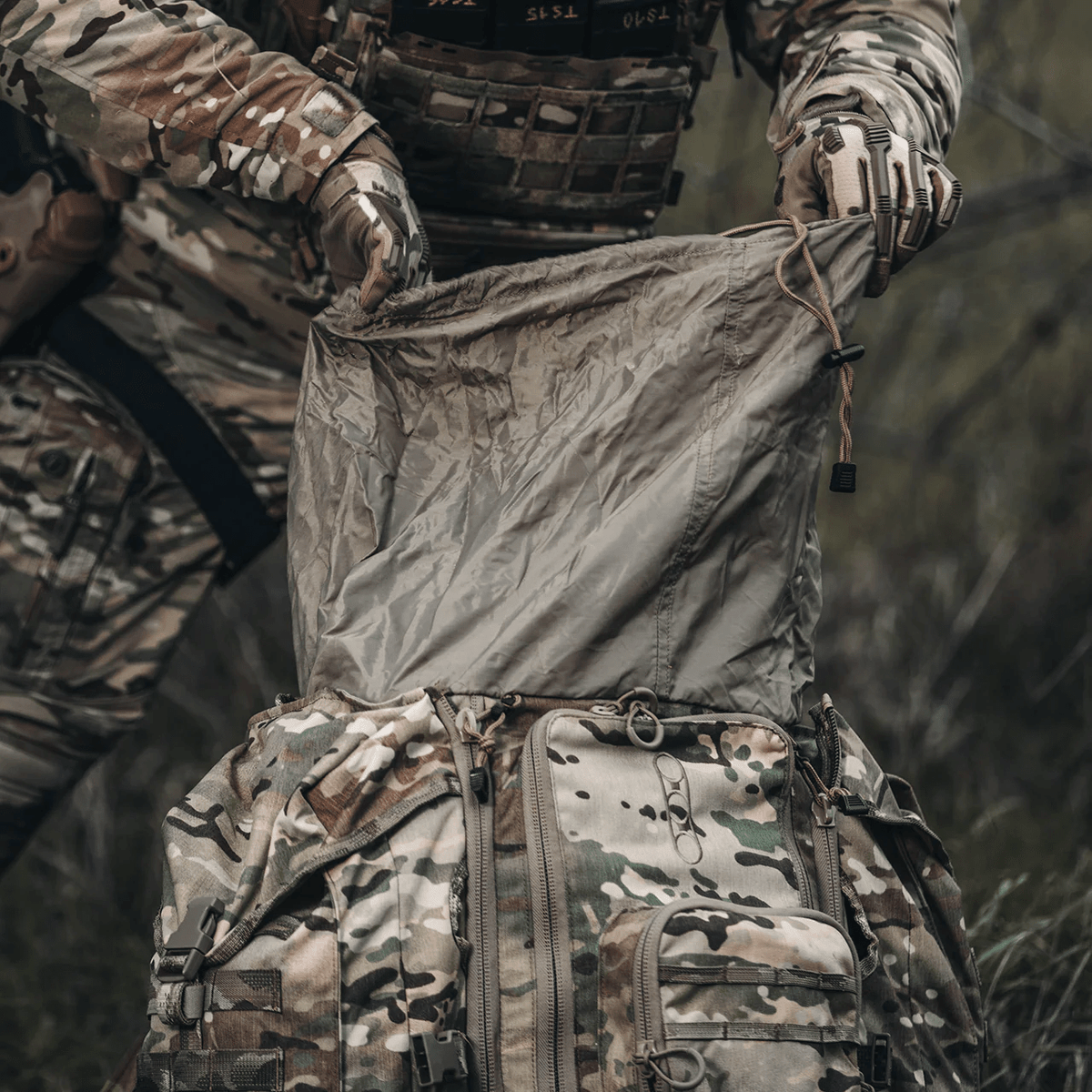 Eine Person in militärischer Tarnkleidung öffnet das obere Fach eines großen EBERLESTOCK G4 Operator Packs, während sie sich im Freien auf einer Wiese befindet.