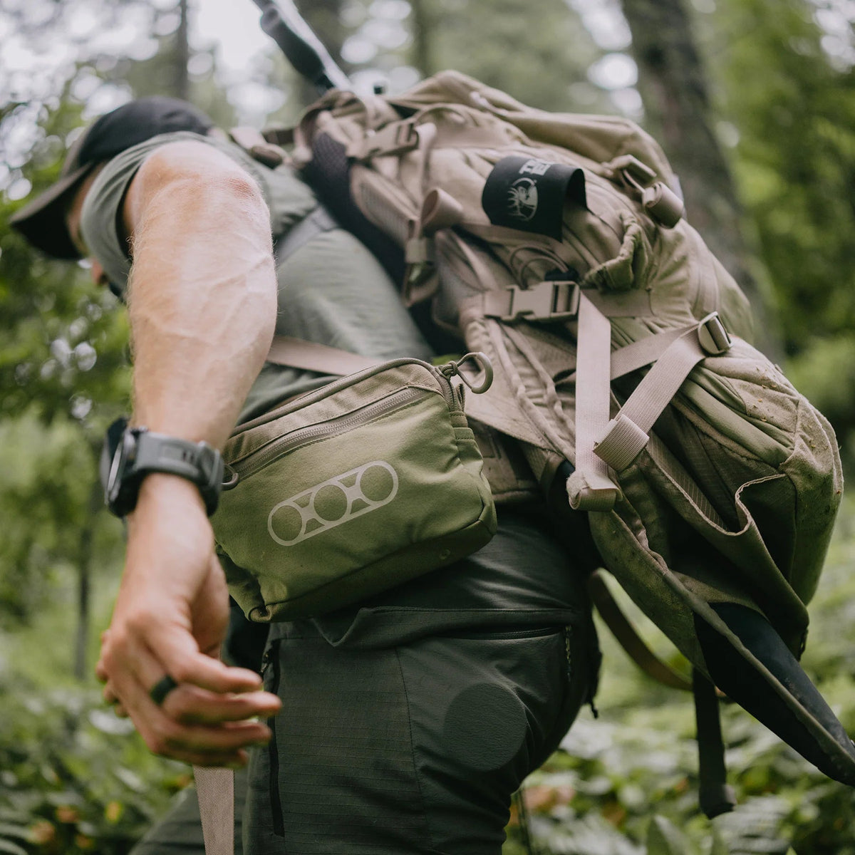 Ein Wanderer mit Outdoor-Ausrüstung und Mütze, der eine EBERLESTOCK General Purpose Pouch Large an seinem Rucksack befestigt hat, wandert durch einen üppigen Wald. Seine MOLLE-kompatible Zusatztasche und seine Uhr sind sichtbar, während er nach vorne greift.