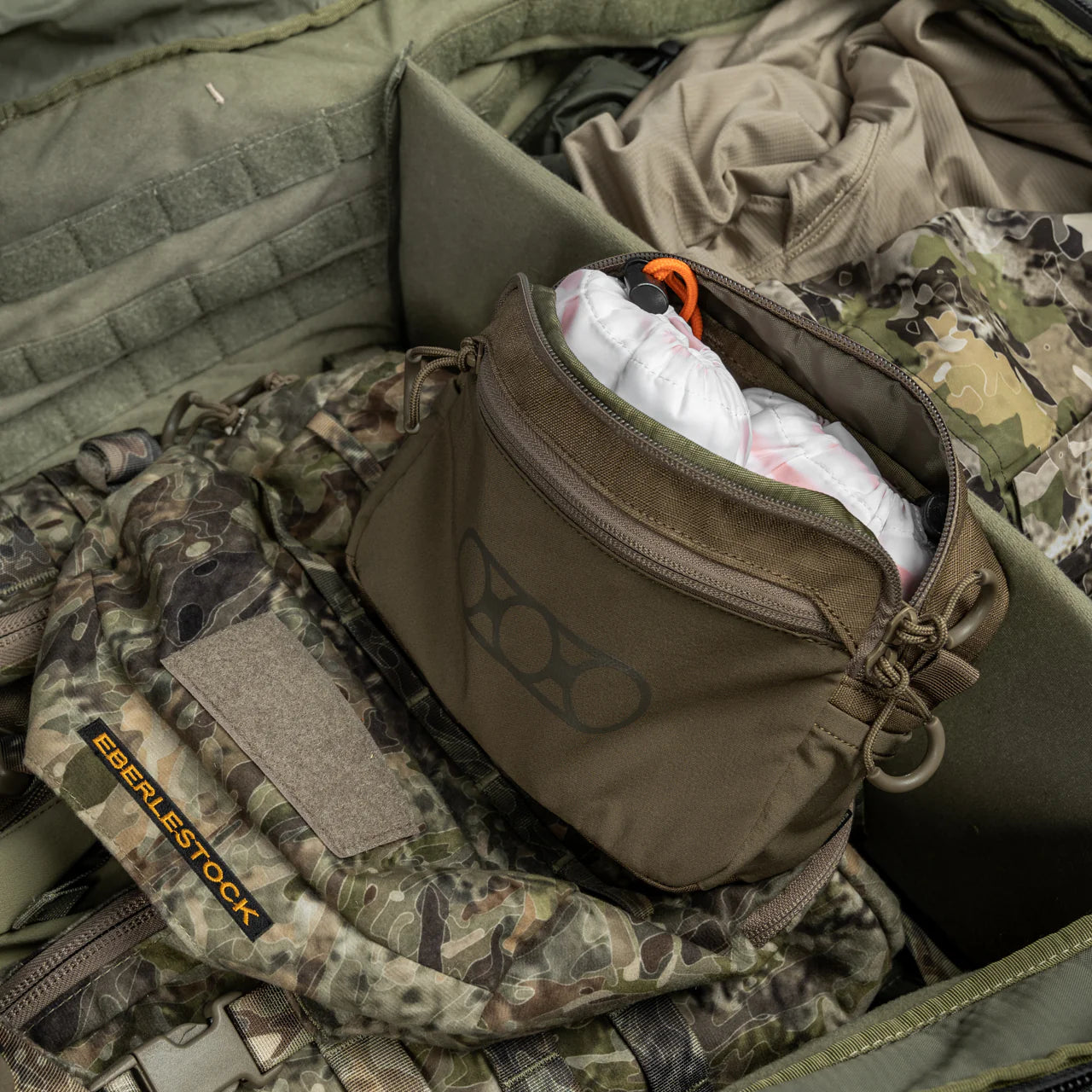 Ein geöffneter Eberlestock Tarnrucksack zeigt eine olivgrüne EBERLESTOCK General Purpose Pouch Large, gefüllt mit weißer und roter Ausrüstung und umgeben von taktischer Kleidung und militärischen Gegenständen.