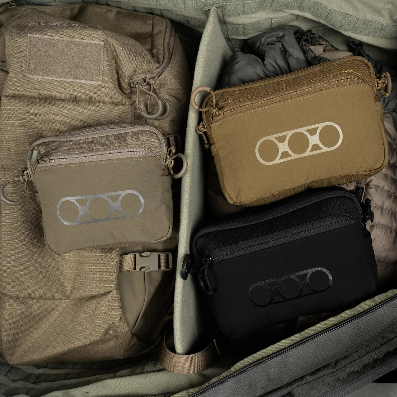 Drei kleine EBERLESTOCK Allzwecktaschen in Hellbraun, Braun und Schwarz - aus strapazierfähigem 500D Cordura-Nylon - sind über der Outdoor-Ausrüstung ausgestellt, darunter ein großer hellbrauner Rucksack und eine Puffjacke, die alle ordentlich in einer offenen Tasche angeordnet sind.