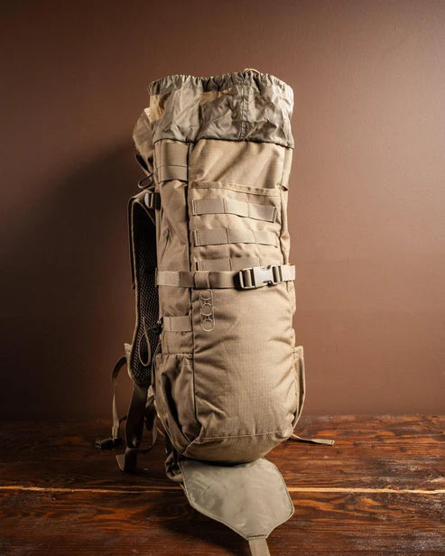 Ein EBERLESTOCK Gunrunner Pack - Mountain in Hellbraun steht aufrecht auf einer hölzernen Unterlage, seine zahlreichen Riemen und Taschen sind vor einem schlichten braunen Hintergrund sichtbar.