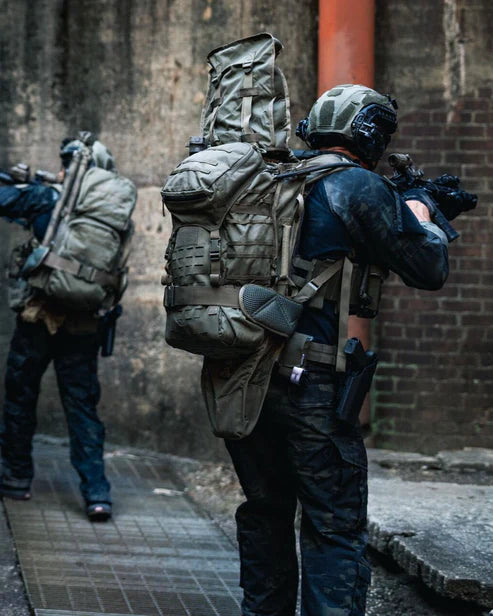 Zwei Personen in taktischer Ausrüstung und mit Helmen, die EBERLESTOCK Gunslinger II - Black Rucksäcke und Gewehre tragen, bewegen sich vorsichtig durch eine verwitterte städtische Gasse mit Ziegel- und Betonflächen.