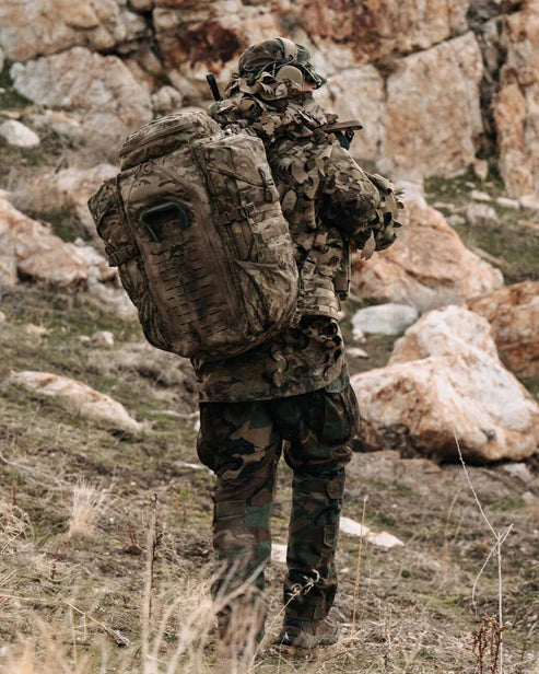 Eine Person in Tarnkleidung und mit einem EBERLESTOCK Halftrack - Black Rucksack geht auf einem grasbewachsenen, felsigen Gelände mit einer Steinmauer im Hintergrund, von der Kamera abgewandt.