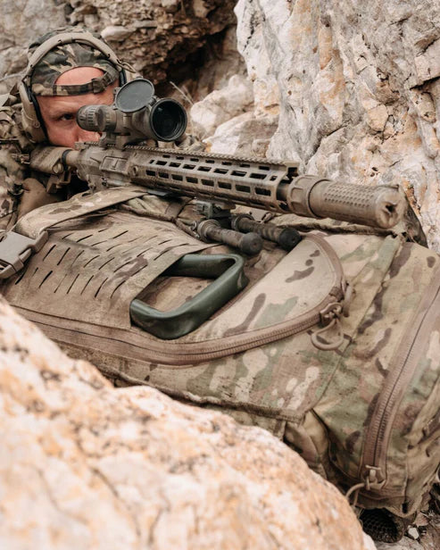 Ein Soldat in Tarnkleidung liegt auf dem Bauch zwischen Felsen und zielt auf ein Scharfschützengewehr mit Zielfernrohr. Er ruht auf einem EBERLESTOCK Halftrack - Dry Earth Rucksack, sein Gesicht ist teilweise sichtbar.