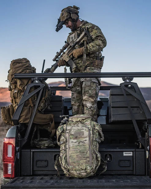 Ein Soldat in Tarnkleidung und mit Helm steht auf der Ladefläche eines Pickups und hält ein Gewehr. An seiner Seite befindet sich der EBERLESTOCK Halftrack-Rucksack in Military Green, umgeben von taktischer Ausrüstung vor dem Hintergrund der Abenddämmerung.