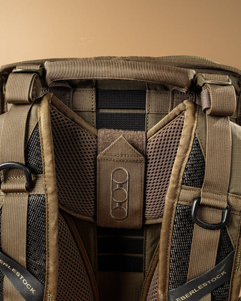 Nahaufnahme des oberen Teils des schwarzen EBERLESTOCK HiSpeed II Technikrucksacks mit gepolsterten Schultergurten, schwarzem Gurtband und Metallringen - ideal für den taktischen Einsatz - vor einem schlichten beigen Hintergrund.