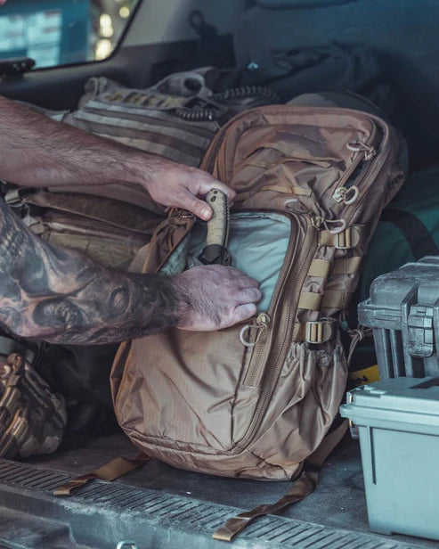 Eine tätowierte Person steckt ein Messer in die Vordertasche eines EBERLESTOCK HiSpeed II - Coyote Brown Rucksacks in einem Fahrzeug, das mit anderen Ausrüstungsgegenständen und Koffern beladen ist.