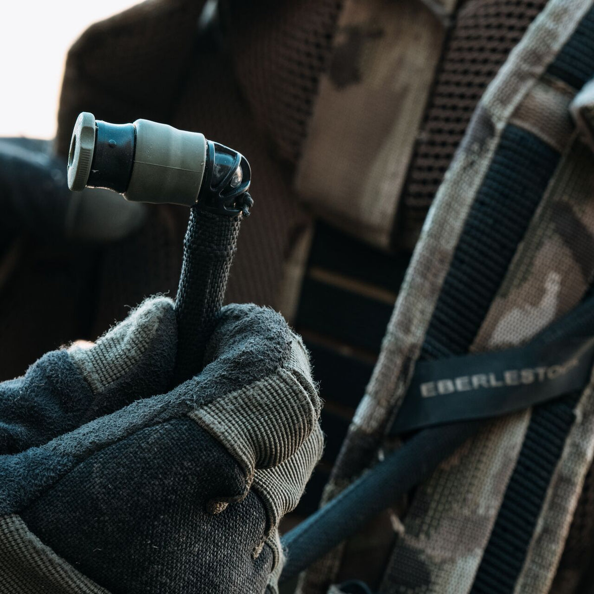 Eine behandschuhte Hand hält das Mundstück eines Eberlestock Hydration System Bladder 3L-Schlauches, der an einem taktischen Rucksack mit EBERLESTOCK-Branding befestigt ist, während sich die Person in Tarnkleidung auf eine zuverlässige Flüssigkeitsversorgung im Freien vorbereitet.