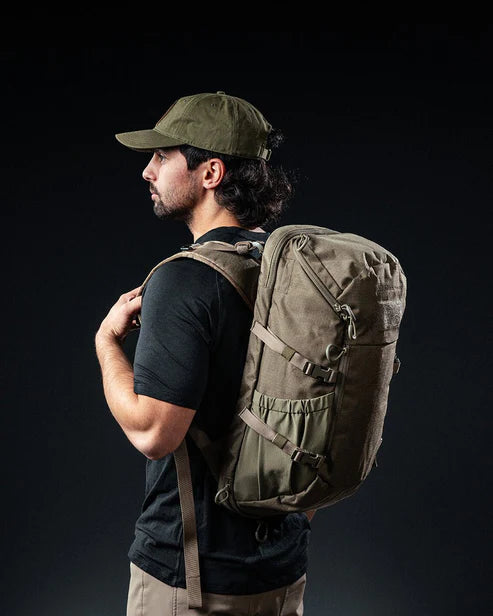 Ein Mann in einem dunklen Hemd, einer khakifarbenen Hose und einer olivgrünen Mütze steht im Profil vor einem schwarzen Hintergrund und trägt den EBERLESTOCK Jacknife - Dry Earth Rucksack auf seinem Rücken.
