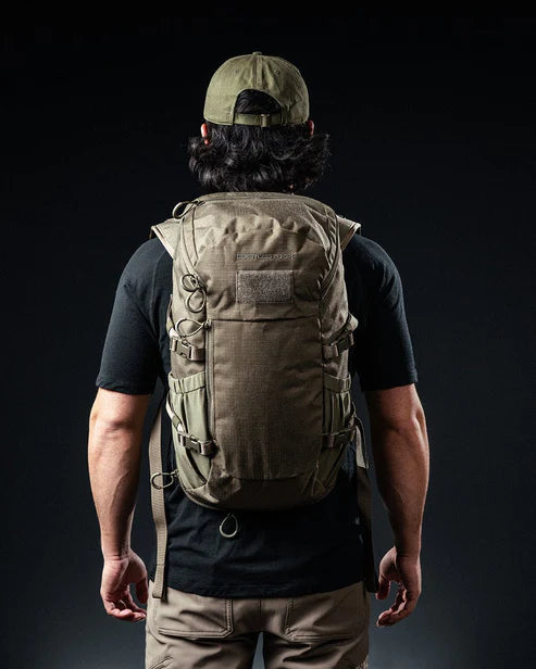 Eine Person mit grüner Mütze, schwarzem T-Shirt und brauner Hose steht mit dem Gesicht zur Wand und präsentiert den EBERLESTOCK Jacknife - Military Green, einen kompakten 26-Liter-Rucksack mit mehreren Riemen und Schlaufen vor einem dunklen Hintergrund.