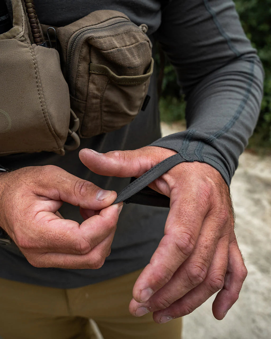 Der EBERLESTOCK Lochsa Zip Merino Hoody in Dark Earth wird von einer Person mit hellbrauner Hose und einer braunen Tasche über der Brust getragen, die ihren Ärmel im Freien inmitten von Grün und einem Feldweg zurechtrückt.