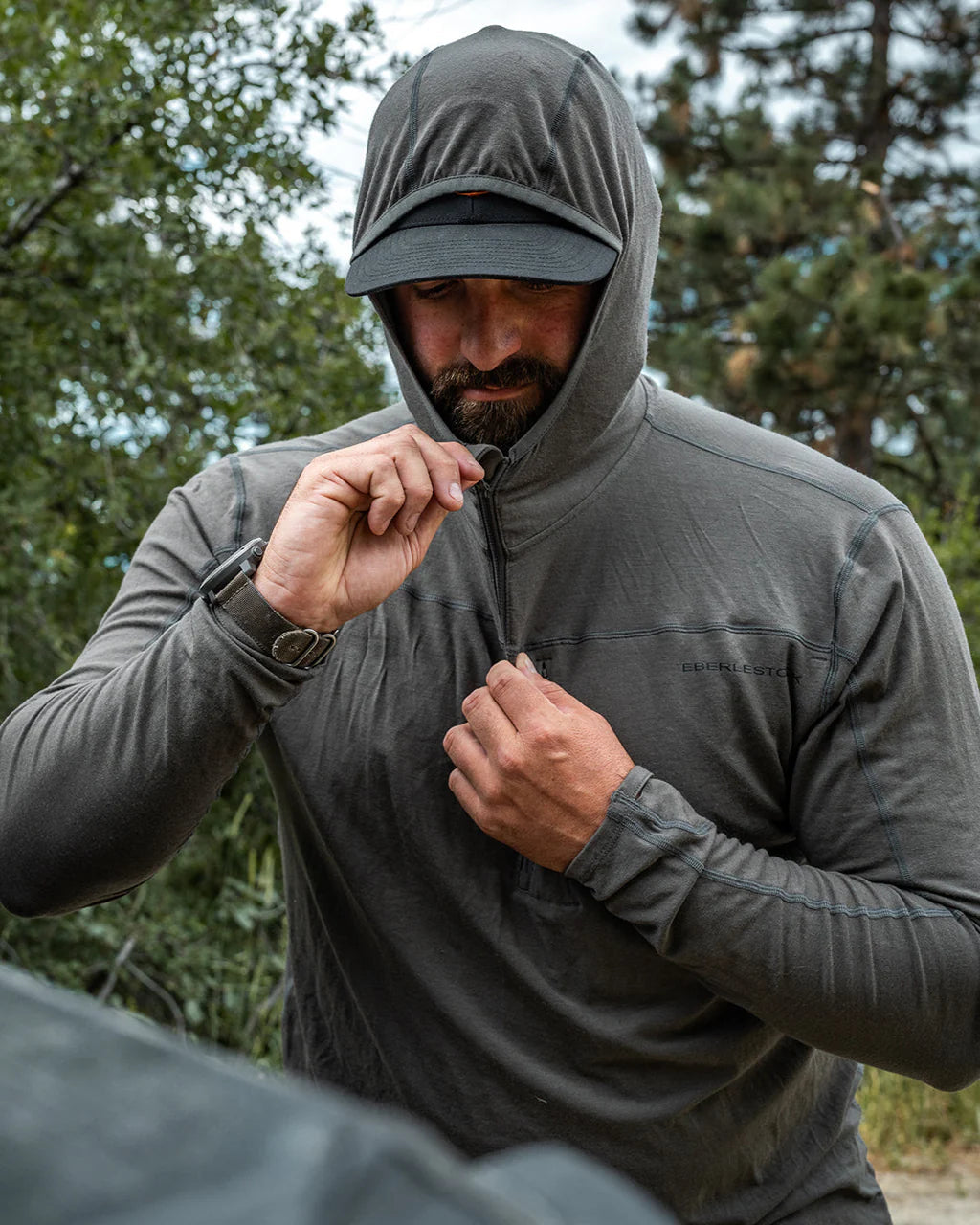 Ein bärtiger Mann im Freien zieht seinen EBERLESTOCK Lochsa Zip Merino Hoody in Loden über eine schwarze Mütze. Er steht inmitten von grünem Laub und Bäumen, während er nach unten schaut, um seine Kapuze anzupassen.