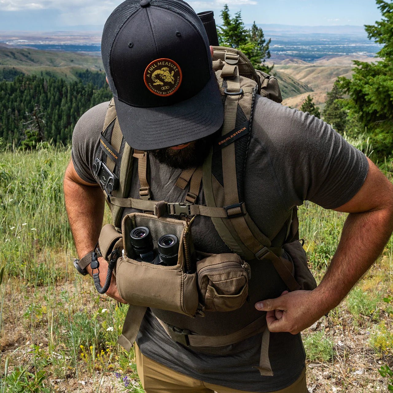 Inmitten von hohem Gras und Bäumen in den Bergen blickt eine Person mit schwarzem Hut, Rucksack und dem EBERLESTOCK Lochsa Merino Short Sleeve - Loden auf ein malerisches Tal im Hintergrund. An ihrem taktischen Gurt hängen Taschen für Ferngläser.