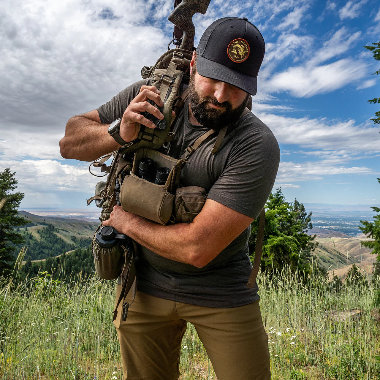 Ein bärtiger Mann, der das EBERLESTOCK Lochsa Merino Short Sleeve - Slate trägt, rückt seinen Rucksack zurecht und hält ein Walkie-Talkie auf einer grasbewachsenen Anhöhe, mit Bäumen, Bergen und einem teilweise bewölkten Himmel im Hintergrund.