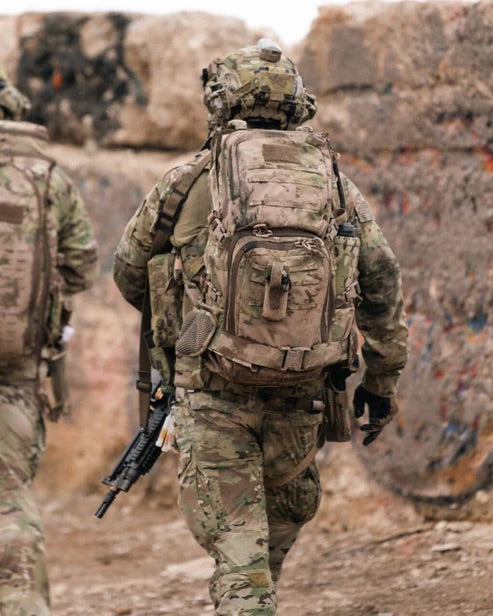 Zwei Soldaten in Tarnkleidung und mit Helmen gehen zwischen Betonbarrieren hindurch, einer trägt einen EBERLESTOCK Lodrag II V2 - Black Rucksack und ein Gewehr, mit dem Rücken zur Kamera in einer rauen Außenumgebung.