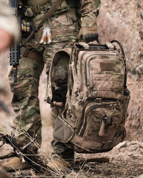 Eine Person in Camouflage steht auf felsigem Gelände und trägt einen EBERLESTOCK Eberlestock Lodrag II V2 - Dry Earth Rucksack mit sichtbarem Gewehr und Munition; ihr Oberkörper und ihre Beine sind zu sehen.