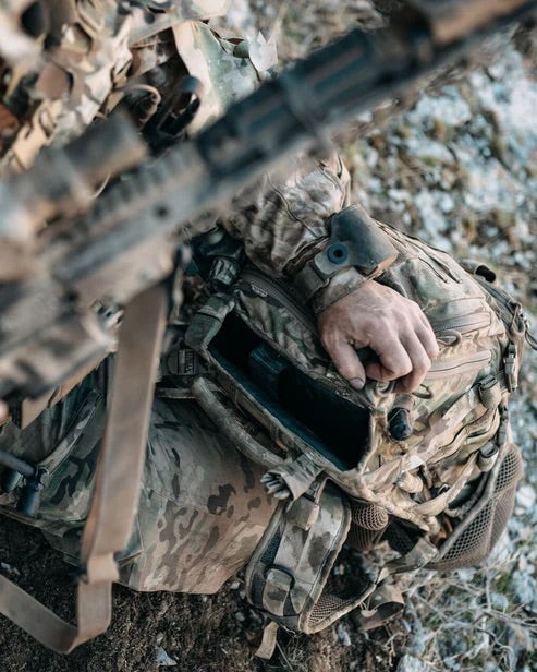 Eine Person in Camouflage kniet auf felsigem Untergrund und greift in einen EBERLESTOCK Eberlestock Lodrag II V2 - Dry Earth Rucksack mit einem teilweise sichtbaren Gewehr, um dessen taktischen Einsatz zu unterstreichen.