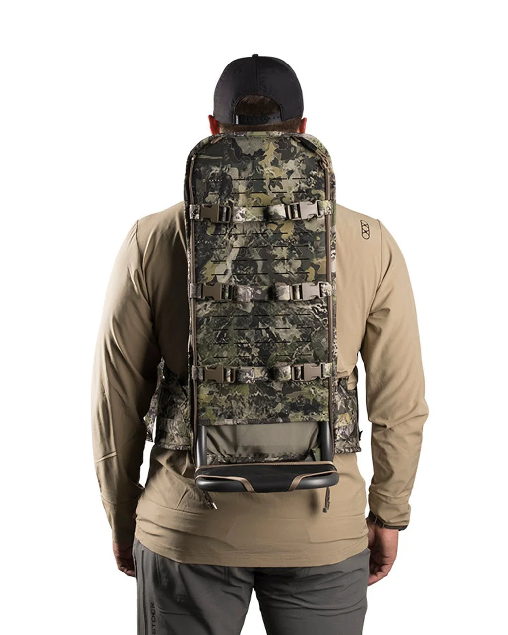 Eine Person, die einen EBERLESTOCK Mainframe - Regular external frame backpack in Camouflage trägt, ist von hinten zu sehen, bekleidet mit einem hellbraunen, langärmeligen Hemd, einer grauen Hose und einer schwarzen Mütze vor einem einfarbig weißen Hintergrund.