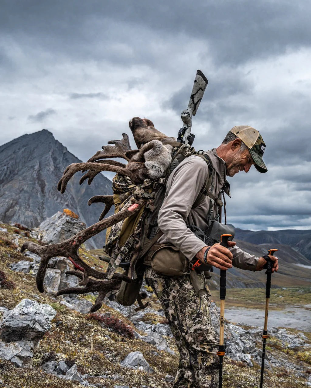 Ein Mann in Camouflage wandert durch zerklüftete Berge und trägt ein Gewehr und ein großes Karibu-Geweih auf seinem EBERLESTOCK Mainframe - Regular Rucksack. Er benutzt Trekkingstöcke und lächelt, mit dramatischen Wolken im Hintergrund.