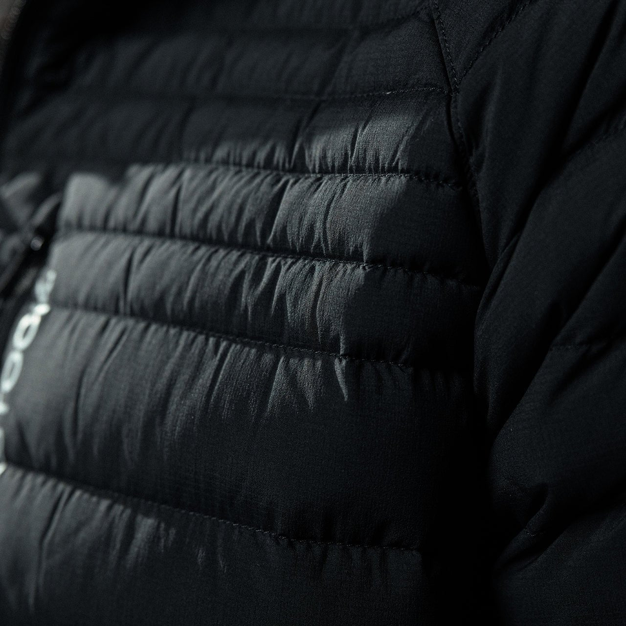 Nahaufnahme des McCall Down Hoody - Black von EBERLESTOCK. Hervorzuheben sind die horizontalen Steppnähte auf dem glänzenden, strukturierten Stoff und das teilweise sichtbare weiße Logo auf der linken Seite.