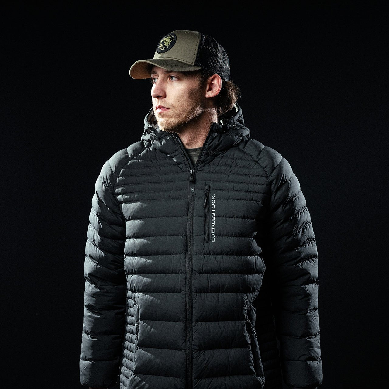 Ein Mann in einem EBERLESTOCK McCall Down Hoody - Black und einer schwarz-olivfarbenen Baseballkappe steht vor einem dunklen Hintergrund und blickt zur Seite.
